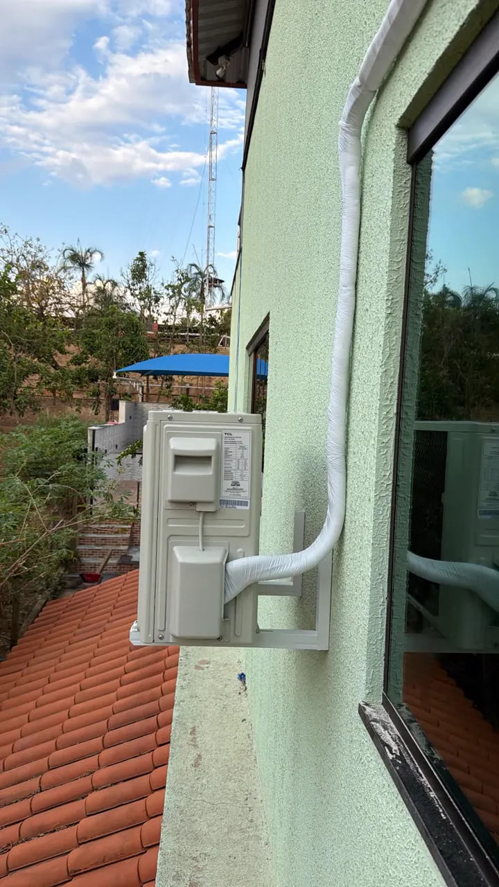 Condensadora em suporte na parede externa verde, com a tubulação numa canaleta branca, acima de um telhado de cerâmica.