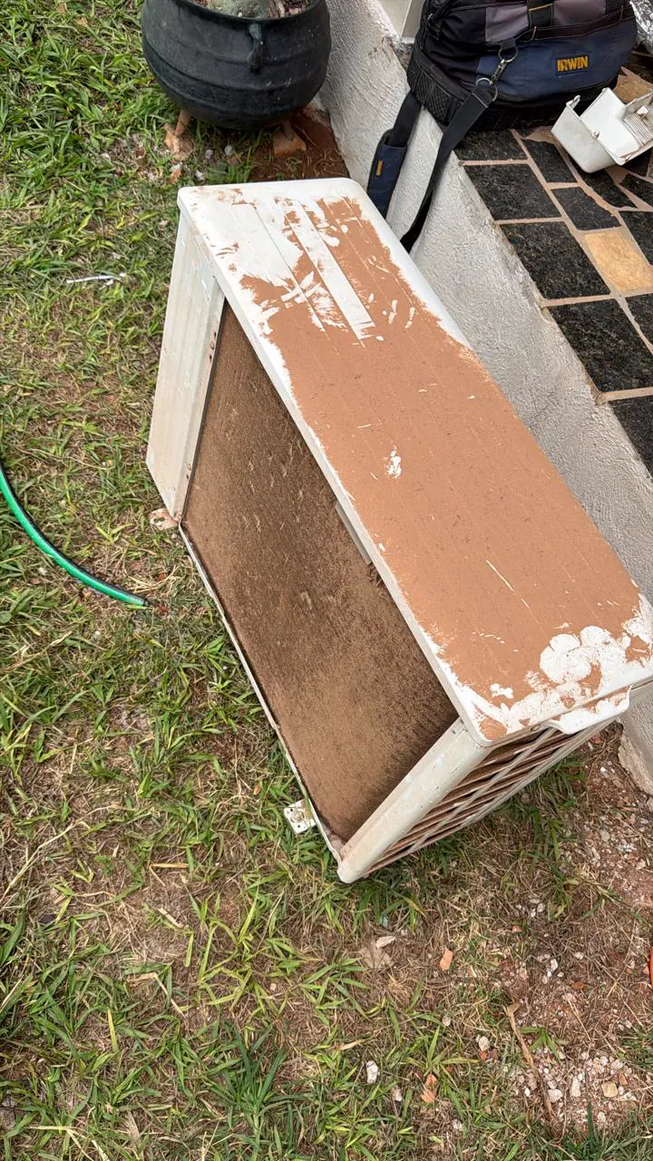 Lateral de uma condensadora com a serpentina marrom de sujeira, apoiada na grama.